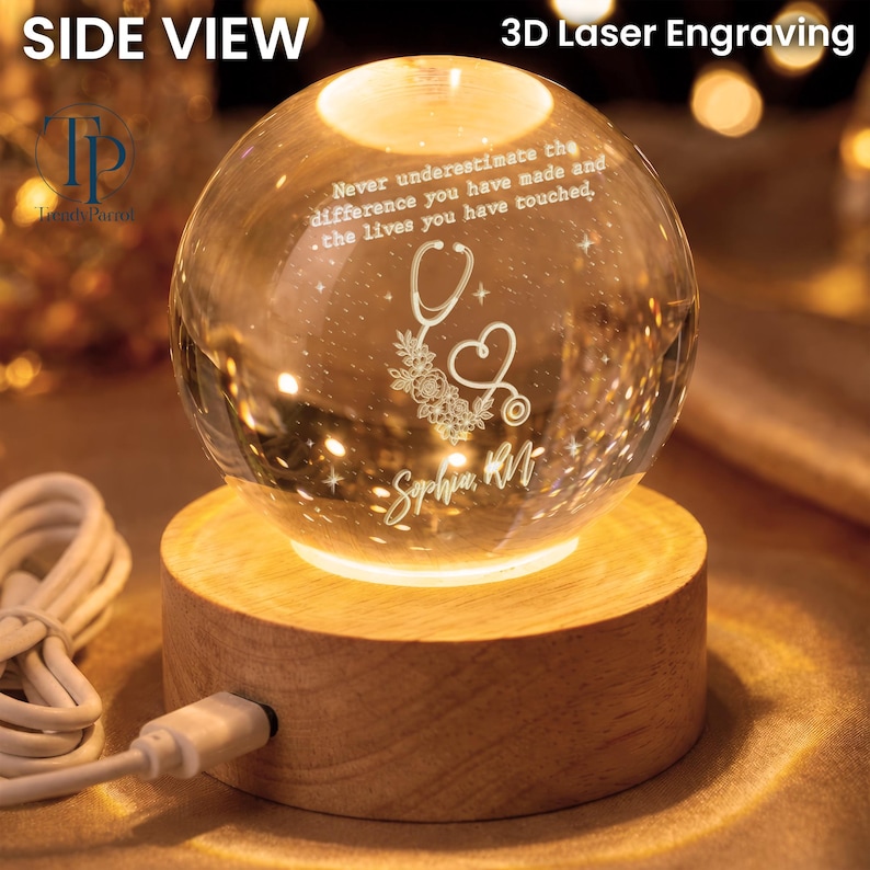 May include: A clear glass sphere with 3D laser engraving resting on a round wooden light base. The engraving features a stethoscope, floral accents, the name Sophia RN, and the text: Never underestimate the difference you have made and the lives you have touched. A white USB power cable is attached to the base.