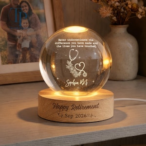 May include: A spherical glass night light on a round wooden base. The glass features an etched stethoscope, floral accents, and the text: Never underestimate the difference you have made and the lives you have touched, Sophia, RN. The base reads: Happy Retirement Sep 2026.