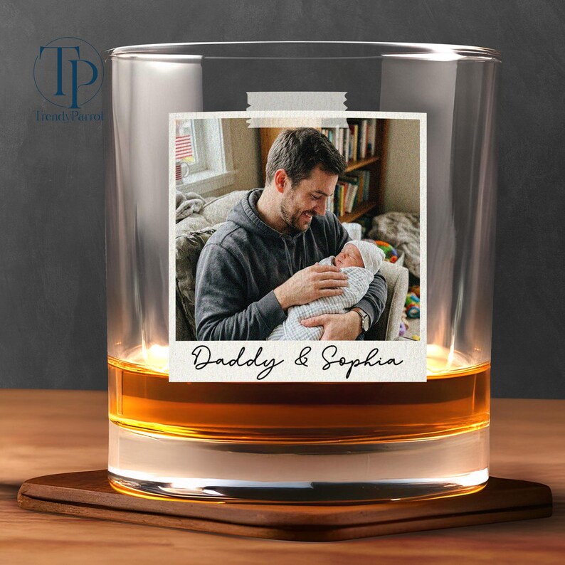 May include: A clear glass tumbler with a photo of a man holding a baby, taped to the glass. The photo has the words "Daddy & Sophia" written on it. The glass is filled with a golden liquid and sits on a wooden coaster.