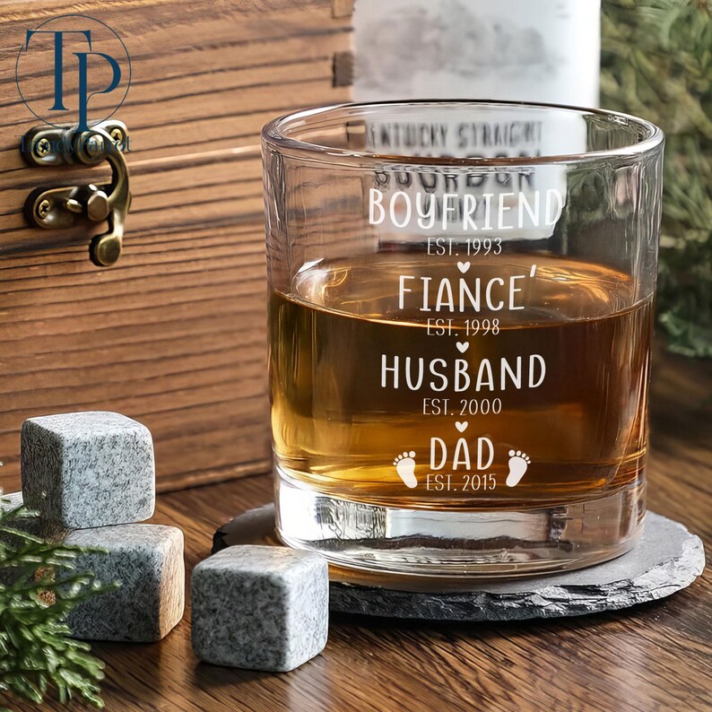 May include: A clear glass whiskey tumbler with the words "Boyfriend Est. 1993", "Fiance' Est. 1998", "Husband Est. 2000", and "Dad Est. 2015" etched on the side. The glass is filled with amber liquid and is sitting on a black slate coaster.