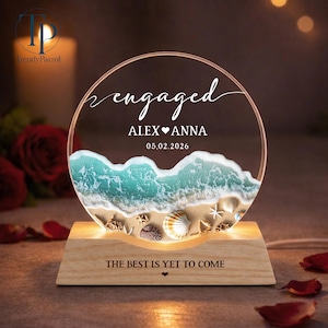 May include: A personalized engagement night light with a beach scene. The clear acrylic circle features the word "engaged", names, and a date. The base is wood, and the text "THE BEST IS YET TO COME" is inscribed on the base.