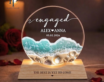 May include: A personalised engagement night light with a beach scene. The clear acrylic circle features the word "engaged", names, and a date. The base is wood, and the text "THE BEST IS YET TO COME" is inscribed on the base.