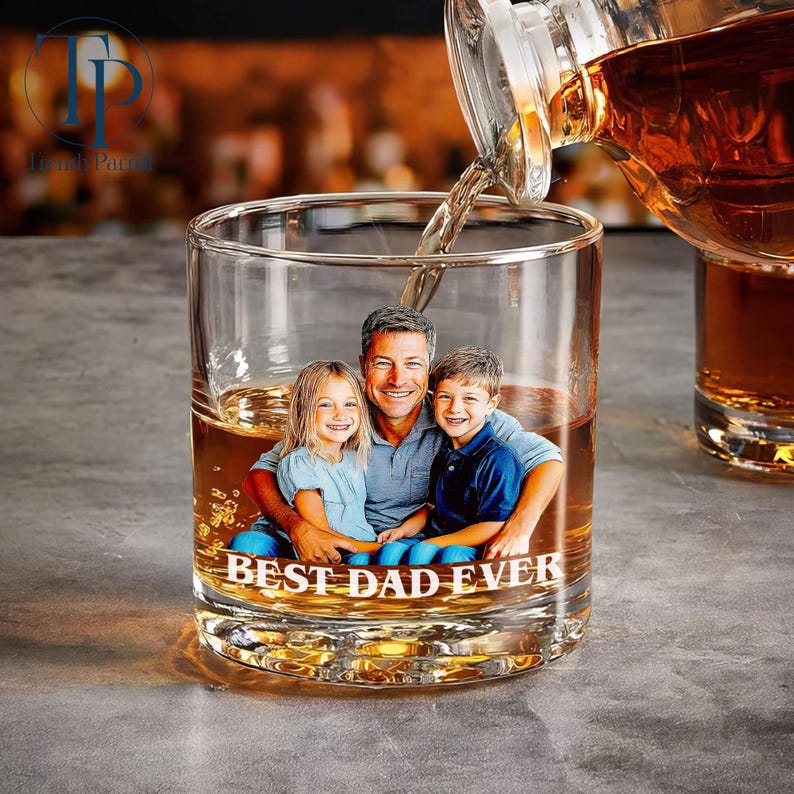 May include: A clear glass tumbler with a photo of a man and two children, with the words "BEST DAD EVER" printed on the bottom. Amber liquid is being poured into the glass from a bottle. The glass is sitting on a gray surface.