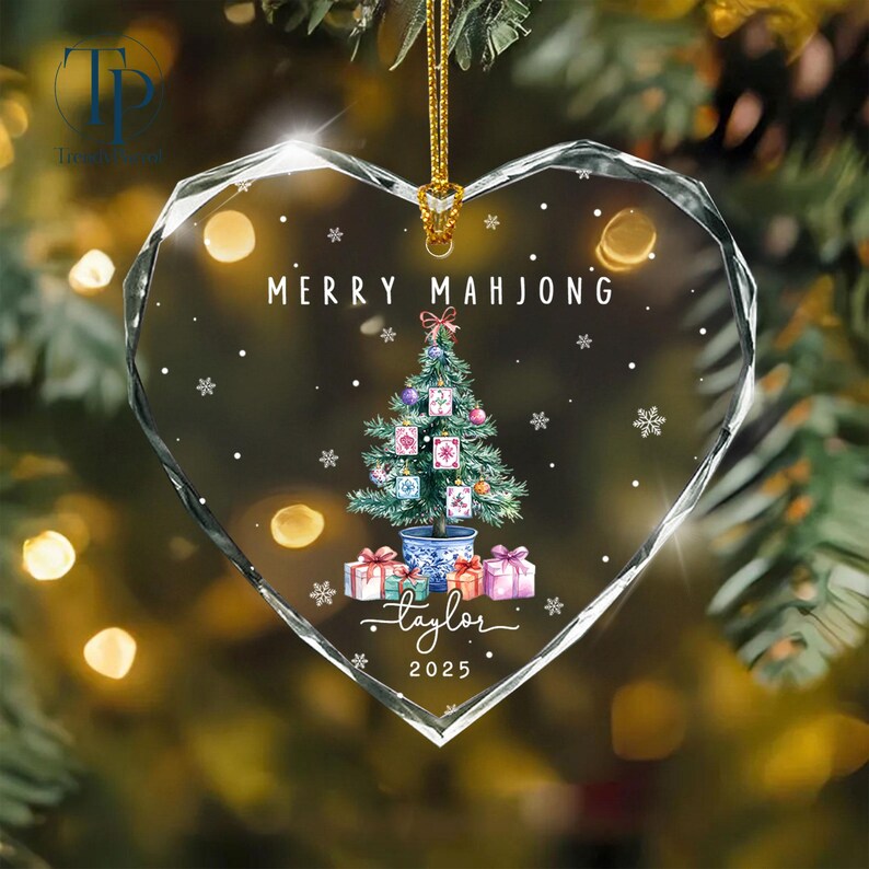 May include: Heart-shaped glass Christmas ornament with the text "MERRY MAHJONG" and a Christmas tree design. The tree is decorated with Mahjong tiles and surrounded by snowflakes. The ornament also includes the name "taylor" and the year "2025".