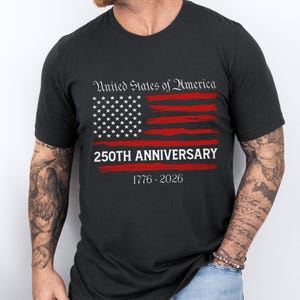 May include: Black t-shirt featuring a distressed American flag design in red and white. The text "United States of America" is above the flag, with "250TH ANNIVERSARY" and "1776-2026" below. A patriotic design for a special occasion.