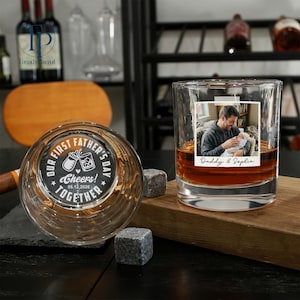 May include: Two personalized whiskey glasses. One glass features a photo of a father and baby with the text "Daddy & Sophia." The other glass has the text "Our First Father's Day! Cheers! Together" and a baby bottle and drink illustration. Includes two square whiskey stones.