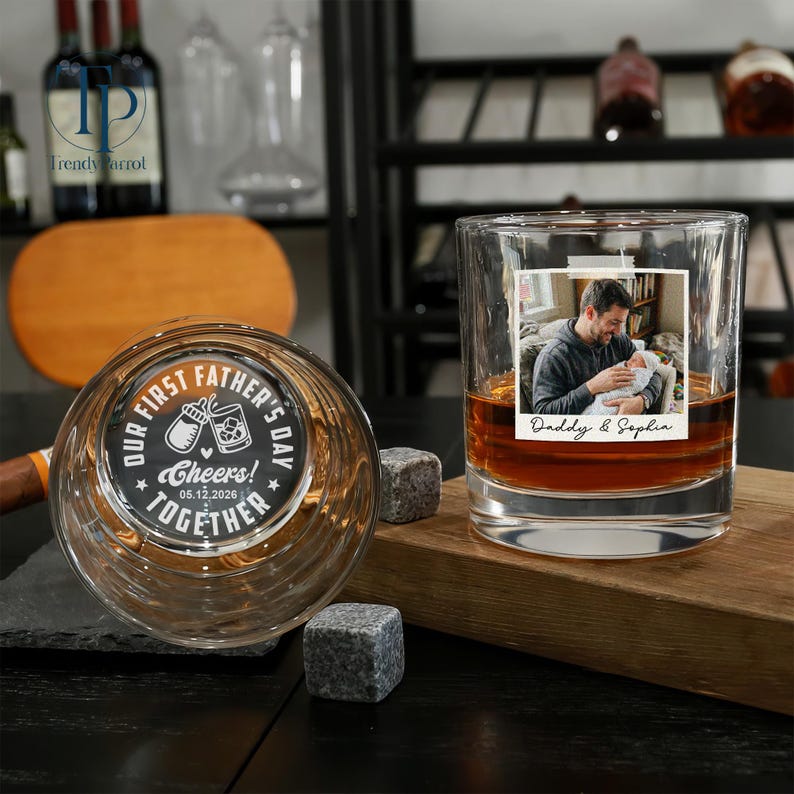 May include: Two personalized whiskey glasses. One glass features a photo of a father and baby with the text "Daddy & Sophia." The other glass has the text "Our First Father's Day! Cheers! Together" and a baby bottle and drink illustration. Includes two square whiskey stones.