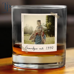May include: A clear glass tumbler with a photo of a man and a child. The photo is taped to the glass and has the words "Grandpa est. 1990". The glass is filled with a brown liquid.