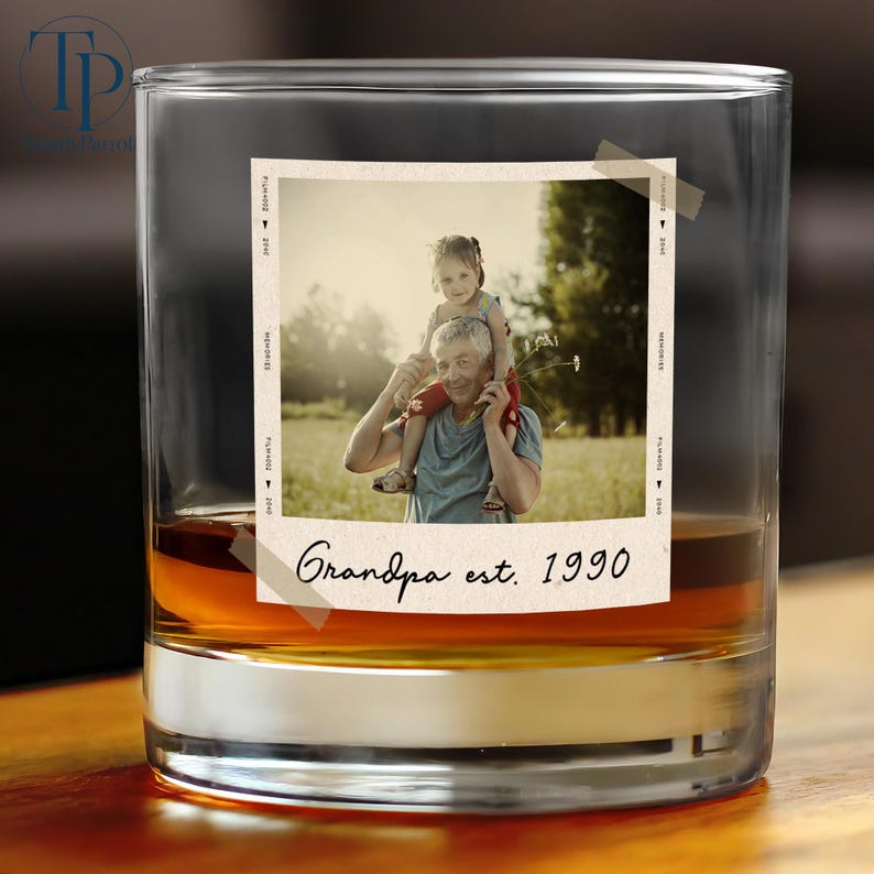 May include: A clear glass tumbler with a photo of a man and a child. The photo is taped to the glass and has the words "Grandpa est. 1990". The glass is filled with a brown liquid.