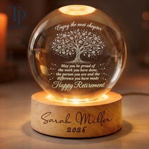 May include: A clear glass sphere with a tree design and the words "Enjoy the next chapter" and "Happy Retirement." It sits on a wooden base engraved with a name and the year 2026. The sphere is illuminated from within.
