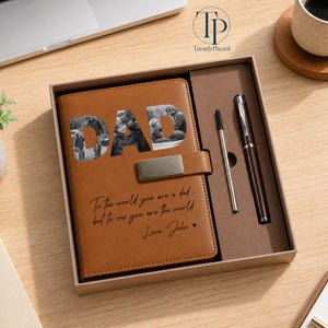 K&ouml;nnte beinhalten: Ein braunes Lederjournal mit dem ausgeschnittenen Wort "DAD", das ein Schwarz-Wei&szlig;-Foto zeigt. Das Journal enth&auml;lt einen Stift und einen Kugelschreiber in einer Geschenkbox. Der Text auf dem Journal lautet: "F&uuml;r die Welt bist du ein Vater, aber f&uuml;r mich bist du die Welt. Liebe, John."