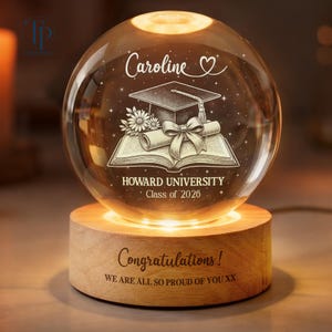 May include: A glowing crystal ball with a wooden base. The ball is engraved with a graduation cap, book, and the text "Caroline", "HOWARD UNIVERSITY Class of 2026". The wooden base reads "Congratulations! WE ARE ALL SO PROUD OF YOU XX".