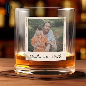 May include: A clear whiskey glass with a photo of a man and a child. The photo is taped to the glass and has the text "Uncle est. 2000". The glass is filled with amber liquid and sits on a wooden coaster.