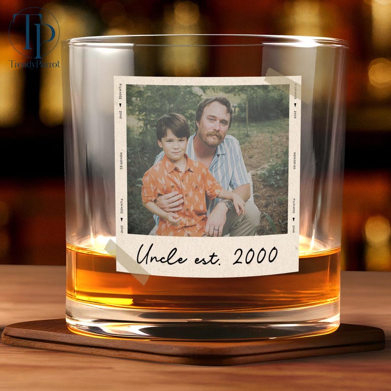 May include: A clear whiskey glass with a photo of a man and a child. The photo is taped to the glass and has the text "Uncle est. 2000". The glass is filled with amber liquid and sits on a wooden coaster.