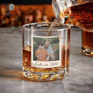 May include: A clear, round whiskey glass with a photo of a man and a child, with the text "Uncle est. 2000". Amber liquid is being poured into the glass from a bottle. The glass sits on a gray surface.