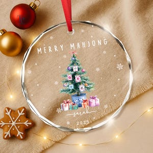 May include: A clear glass Christmas ornament with a red ribbon. The ornament features a watercolor Christmas tree with Mahjong tiles, gifts, and the text "MERRY MAHJONG" and "Sarah 2025". Two Christmas balls and a gingerbread cookie are also visible.