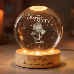 May include: A clear glass sphere with the words "Chapter SIXTY" and "Sarah" engraved, along with a floral design. The sphere sits on a wooden base that reads "Every chapter brings new growth."