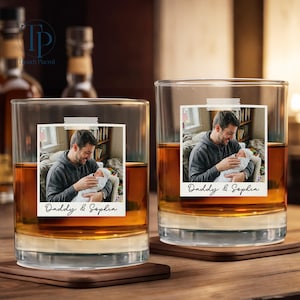 May include: Two clear whiskey glasses with a photo of a man holding a baby, labeled "Daddy & Sophia." The glasses sit on wooden coasters, filled with amber liquid. The background includes bottles and a wooden surface.