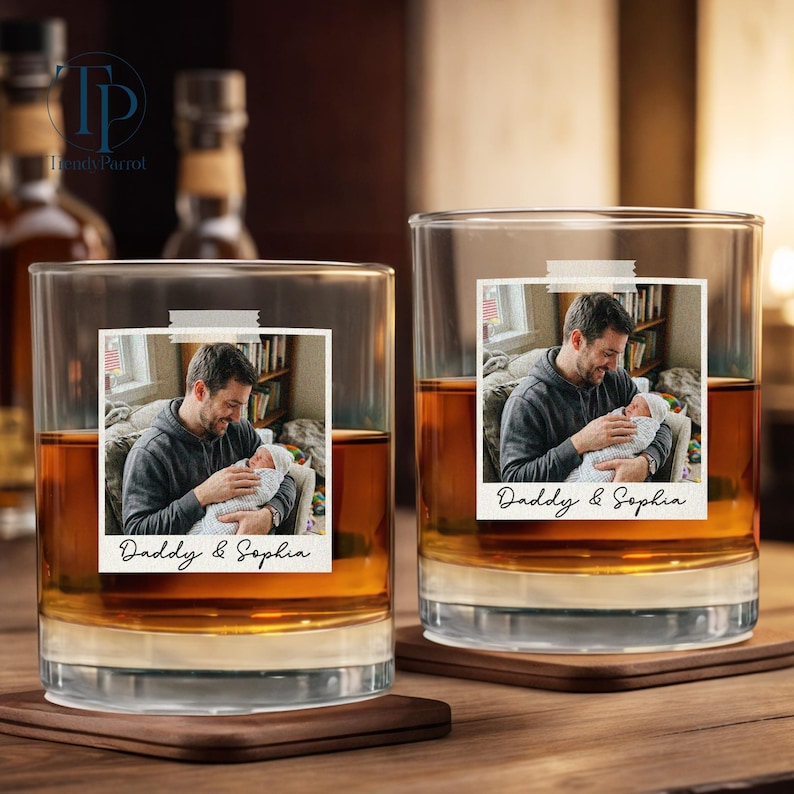 May include: Two clear whiskey glasses with a photo of a man holding a baby, labeled "Daddy & Sophia." The glasses sit on wooden coasters, filled with amber liquid. The background includes bottles and a wooden surface.