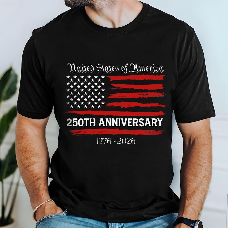 May include: Black t-shirt featuring a distressed American flag design with white stars and red stripes. The text "United States of America" is above the flag, with "250TH ANNIVERSARY" and the years "1776-2026" below.