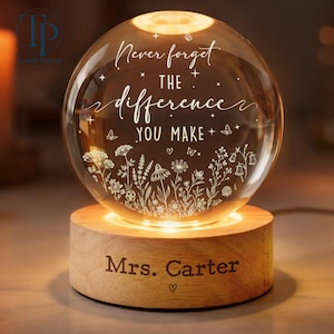May include: A spherical glass night light on a cylindrical wooden base. The glass sphere features the engraved text "Never forget THE difference YOU MAKE" surrounded by small stars, butterflies, and wildflowers. The wooden base is engraved "Mrs. Carter".