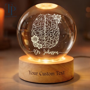 May include: A spherical glass lamp featuring a brain design and floral details, lit from within. The lamp rests on a wooden base, with the text "Your Custom Text" and the name "Dr. Johnson" engraved.