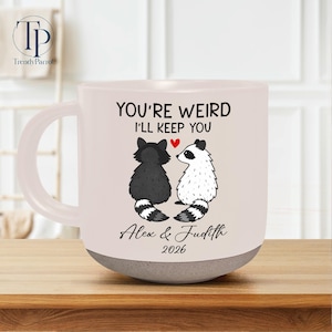 May include: White ceramic mug with a gray base. The mug features a black and white raccoon couple illustration with the text "YOU'RE WEIRD I'LL KEEP YOU" and names "Alex & Judith 2026".