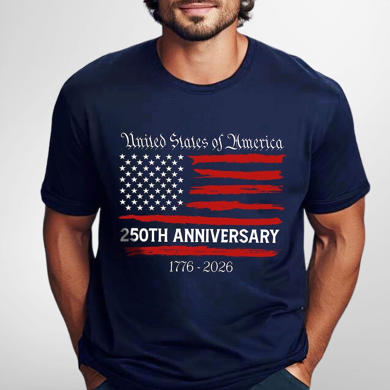 May include: Navy blue t-shirt with a distressed American flag design in red and white. The shirt features the text "United States of America" above the flag and "250th Anniversary 1776-2026" below it. A patriotic themed tee.
