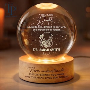 May include: A glowing glass sphere with the text "A Truly Great Doctor" and a stethoscope design, personalized with "Dr. Sarah Smith." It sits on a wooden base with the inscription "Never underestimate the difference you make."