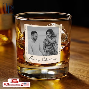 May include: A rocks glass filled with amber liquid and ice cubes. A photo with a couple is adhered to the glass, with the words "Be my Valentine" below the photo. The glass sits on a wooden surface.