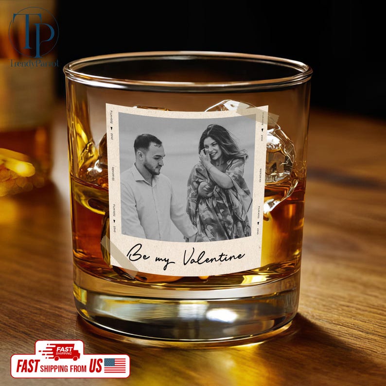 May include: A rocks glass filled with amber liquid and ice cubes. A photo with a couple is adhered to the glass, with the words "Be my Valentine" below the photo. The glass sits on a wooden surface.