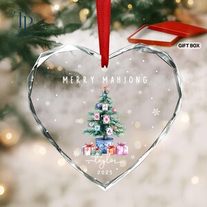 May include: Heart-shaped glass Christmas ornament with a red ribbon. The ornament features a Christmas tree decorated with mahjong tiles, gifts, and the text "MERRY MAHJONG" and "Taylor 2025". Includes a red gift box.