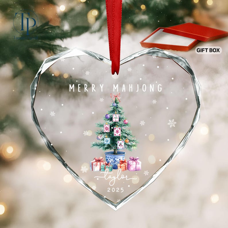 May include: Heart-shaped glass Christmas ornament with a red ribbon. The ornament features a Christmas tree decorated with mahjong tiles, gifts, and the text "MERRY MAHJONG" and "Taylor 2025". Includes a red gift box.
