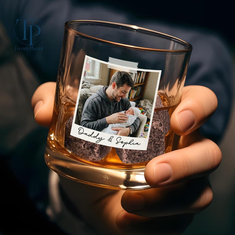 May include: A clear whiskey glass with a photo of a man holding a baby, taped to the front. The photo has the words "Daddy & Sophia" written on it. The glass is filled with brown ice cubes and held by a person.