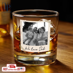 May include: A clear, lowball whiskey glass filled with amber liquid and ice cubes. A black and white photo of a family is adhered to the glass, with the text "We love Dad" below the photo. The glass sits on a wooden surface.