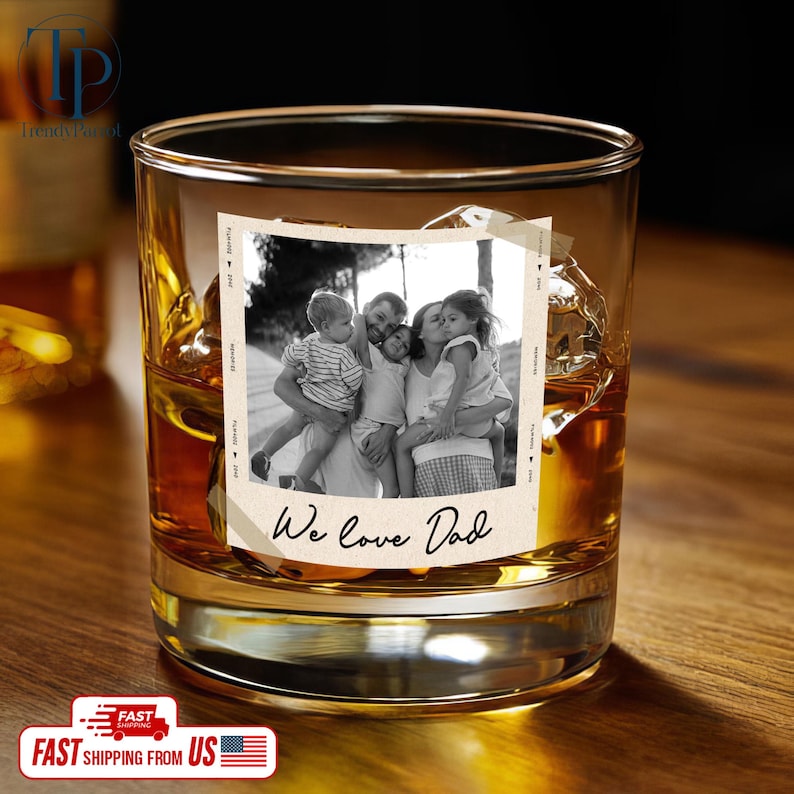 May include: A clear, lowball whiskey glass filled with amber liquid and ice cubes. A black and white photo of a family is adhered to the glass, with the text "We love Dad" below the photo. The glass sits on a wooden surface.