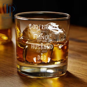 May include: A clear glass tumbler with the words "Boyfriend Est. 1993", "Fiance Est. 1998", "Husband Est. 2000", and "Dad Est. 2015" etched on the side. The glass is filled with amber liquid and ice cubes.