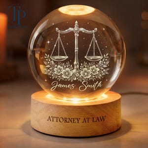 May include: A clear glass sphere featuring a scale of justice design, floral details, and the name "James Smith." It rests on a wooden base engraved with "ATTORNEY AT LAW." The sphere is lit with a warm, inviting light.