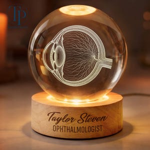 May include: A clear glass sphere lamp featuring an internal 3D laser-etched anatomical diagram of a human eye. The sphere rests on a circular light-up wooden base engraved with the text Taylor Steven OPHTHALMOLOGIST in a dark brown font.