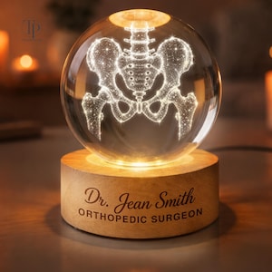May include: A clear glass sphere featuring an internal laser-etched human pelvic bone structure, illuminated from below. The sphere rests on a cylindrical wooden base engraved with the text Dr. Jean Smith ORTHOPEDIC SURGEON in black lettering.