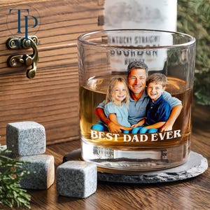 May include: A clear whiskey glass with a photo of a man and two children, with the text "BEST DAD EVER." The glass sits on a coaster, with three gray stone cubes nearby. A wooden box is in the background.