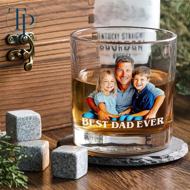 May include: A clear whiskey glass with a photo of a man and two children, with the text "BEST DAD EVER." The glass sits on a coaster, with three gray stone cubes nearby. A wooden box is in the background.