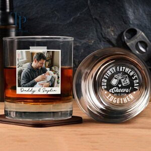 May include: A personalized whiskey glass with a photo of a person holding a baby, labeled "Daddy & Sophia." The glass is filled with amber liquid and rests on a wooden coaster. The bottom of the glass has the text "OUR FIRST FATHER'S DAY TOGETHER Cheers! 05.12.2026."