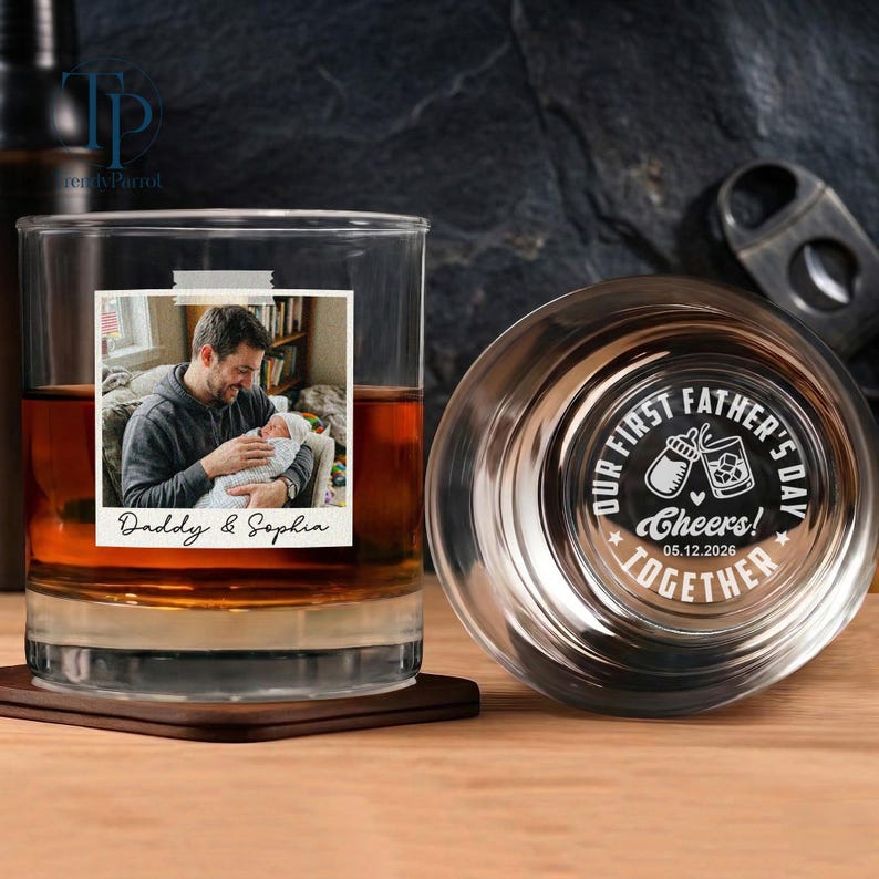 May include: A personalized whiskey glass with a photo of a person holding a baby, labeled "Daddy & Sophia." The glass is filled with amber liquid and rests on a wooden coaster. The bottom of the glass has the text "OUR FIRST FATHER'S DAY TOGETHER Cheers! 05.12.2026."