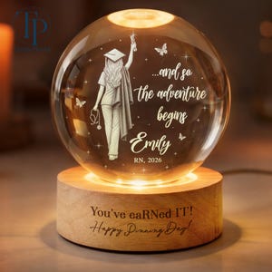 May include: A lighted crystal ball with a nurse illustration and the text "...and so the adventure begins Emily RN, 2026". The wooden base reads "You've eaRNed IT! Happy Pinning Day!"