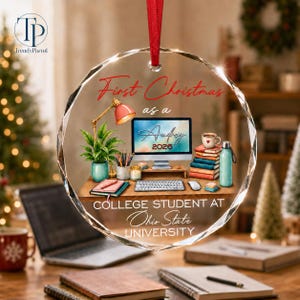 May include: A round, clear glass ornament with a red ribbon, featuring a printed desk scene with a computer monitor displaying 'Audrey 2026', a lamp, books, a water bottle, and the text 'First Christmas as a COLLEGE STUDENT AT Ohio State UNIVERSITY'.