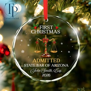 May include: A clear, circular glass Christmas ornament featuring a gold scale of justice graphic, red holly accents, and white text reading FIRST CHRISTMAS ADMITTED STATE BAR OF ARIZONA John Smith, Esq. 2026, suspended by a red ribbon.