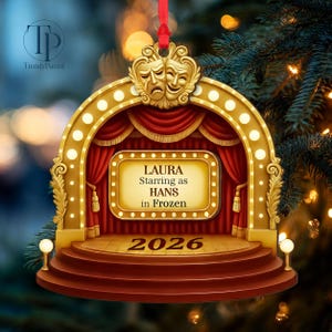 May include: A gold-toned theater stage ornament featuring red curtains, a marquee with the text LAURA Starring as HANS in Frozen, and the year 2026 on the steps. The top displays comedy and tragedy masks, and a red ribbon is attached for hanging.