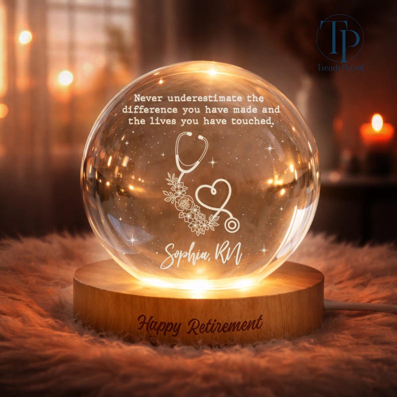 May include: A clear glass sphere night light on a round wooden base. The sphere features the engraved text: Never underestimate the difference you have made and the lives you have touched, a stethoscope heart design, and the name Sophia, RN. Happy Retirement.