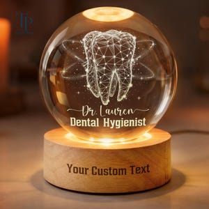 May include: A lit dental-themed crystal ball lamp. The clear sphere has a detailed tooth design with the text "Dr. Lauren Dental Hygienist". The wooden base has the text "Your Custom Text".